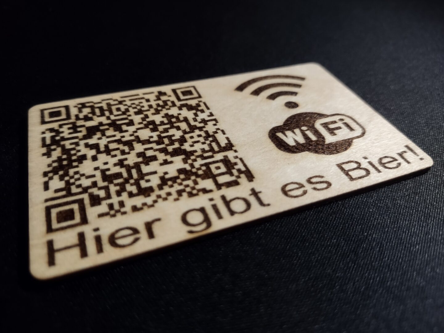 Eine Visitenkarte aus Holz, auf der ein QR-Code und das WiFi-Logo abgebildet sind. Zusätzlich steht die SSID "Hier gibt es Bier" am unteren Rand